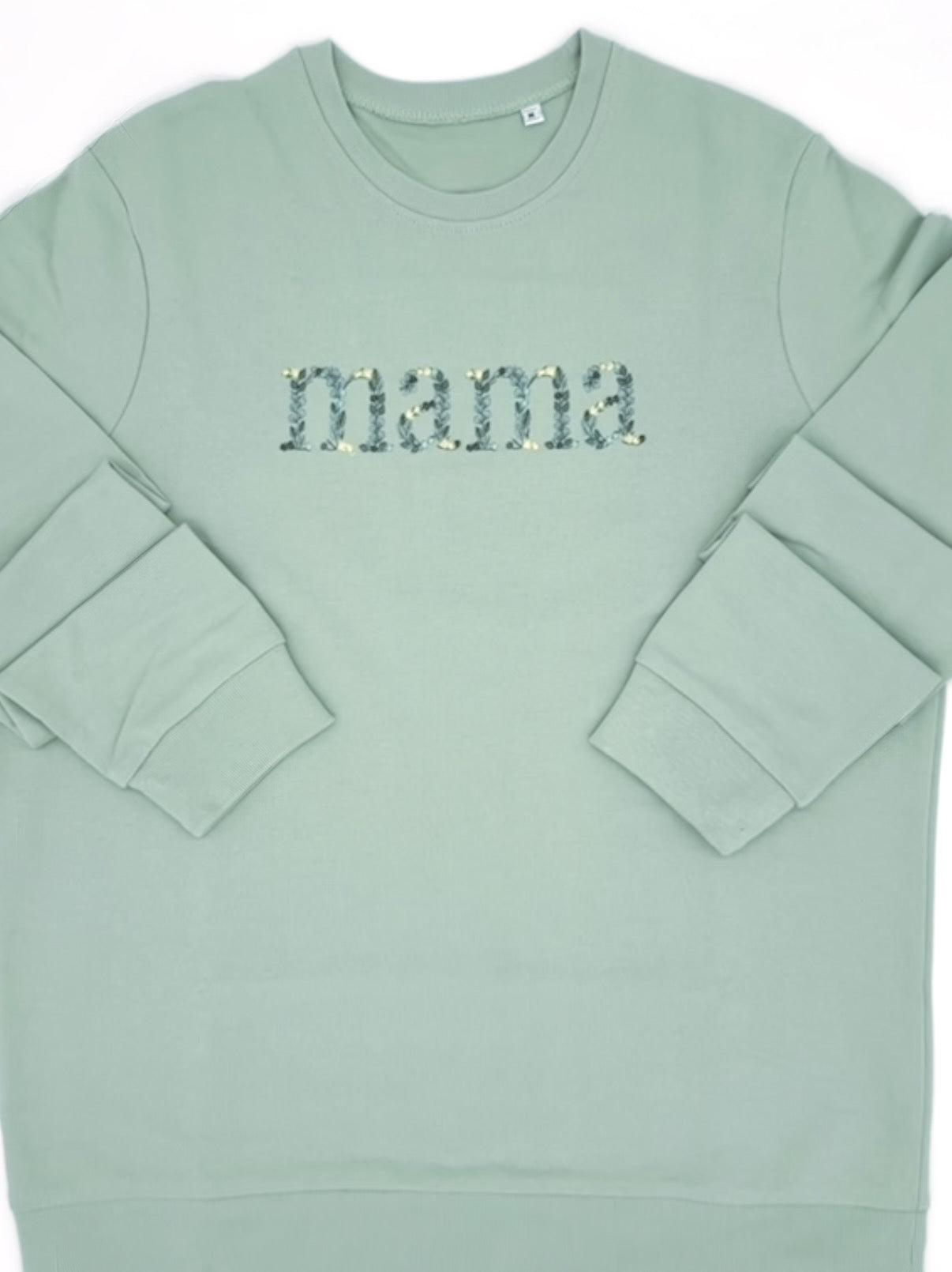 Stickerei auf der Brust des Wortes "Mama". Die einzelnen Buchstaben sind in Form von Blättern in verschiedenen Gruentoenen und gelben Blüten bestickt.
