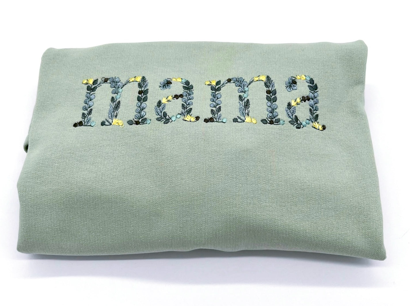 Detailansicht der Stickerei "Mama". Die einzelnen Buchstaben sind in Form von Blättern in verschiedenen Gruentoenen und gelben Blüten bestickt.