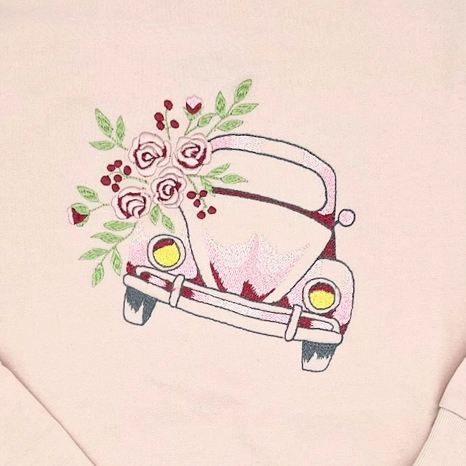 Detailansicht des gestickten VW Kaefer Oldtimer. Links vom Kaefer ist ein Inset Blumen. Die Stickerei ist in Rosa-Rot-Toenen, gruen im Blumenstrauss und gelben Schweinwerfern.
