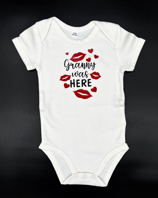 Baby Kurzarm-Body in weiss. Ueber die gesamte Brust sind Kussmuender / Lippenabdruecke und Herzen in rot abgebildet. Dazwischen steht kursiv in schwarz "Granny was" und in Blockschrift "HERE" in schwarz.
