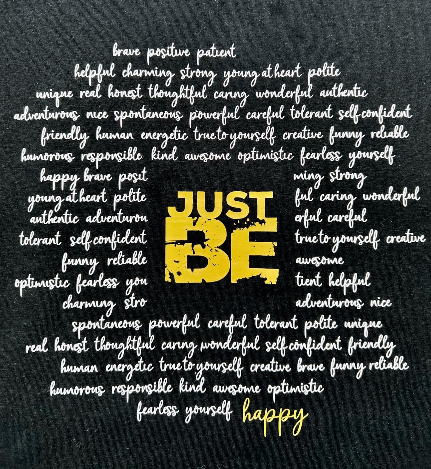 T-Shirt in schwarz. Ueber die gesamt Brust sind viele Woerter kursiv in weiss geschrieben die einen Kreis formen. Im Zentrum steht in Blockschrift in gold "JUST BE" und schliesst den Kreis der Woerter unten rechts mit dem Wort "happy" in gold.