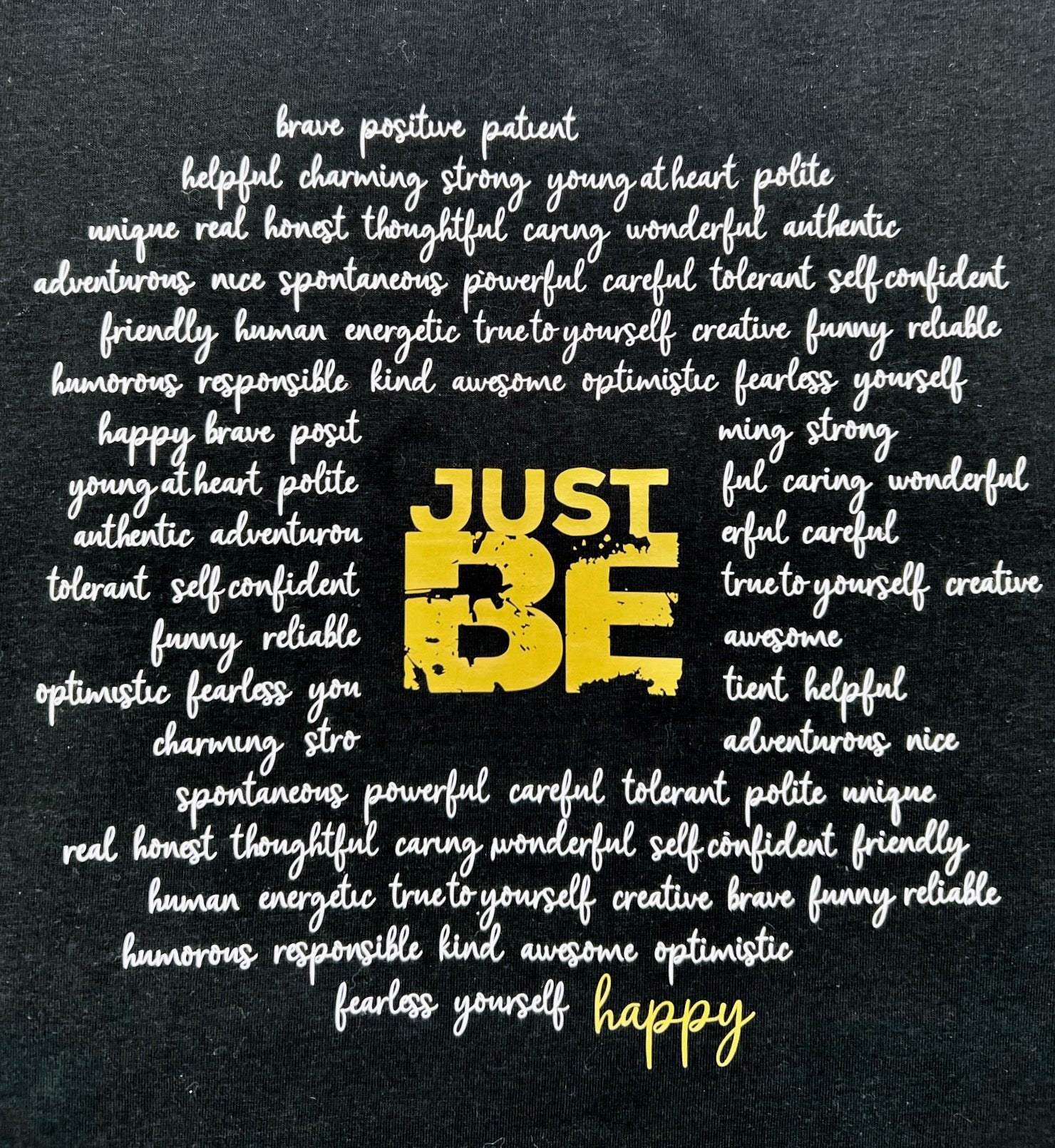 T-Shirt in schwarz. Ueber die gesamt Brust sind viele Woerter kursiv in weiss geschrieben die einen Kreis formen. Im Zentrum steht in Blockschrift in gold "JUST BE" und schliesst den Kreis der Woerter unten rechts mit dem Wort "happy" in gold.
