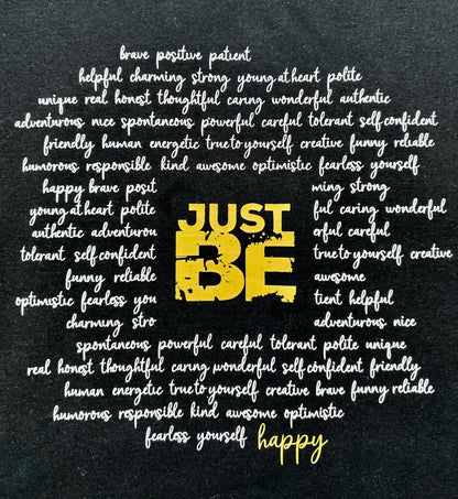T-Shirt in schwarz. Ueber die gesamt Brust sind viele Woerter kursiv in weiss geschrieben die einen Kreis formen. Im Zentrum steht in Blockschrift in gold "JUST BE" und schliesst den Kreis der Woerter unten rechts mit dem Wort "happy" in gold.