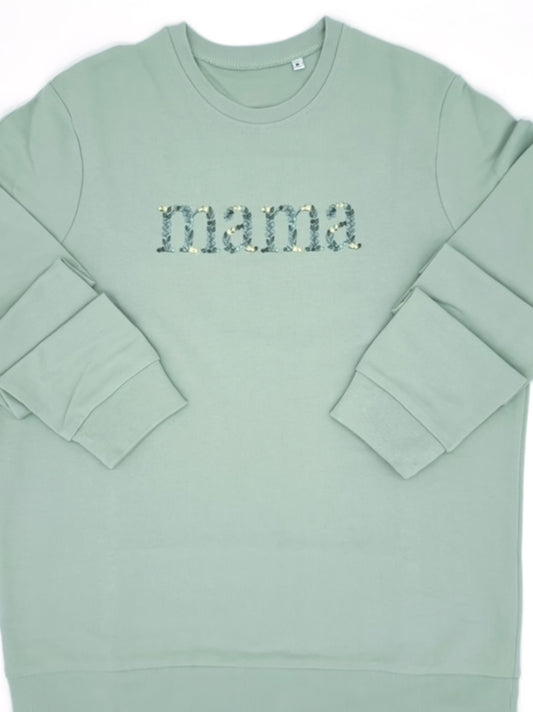 Stickerei auf der Brust des Wortes "Mama". Die einzelnen Buchstaben sind in Form von Blättern in verschiedenen Gruentoenen und gelben Blüten bestickt.