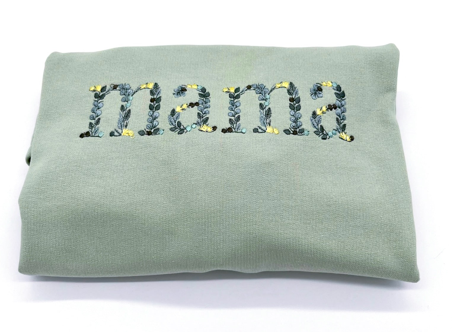 Detailansicht der Stickerei "Mama". Die einzelnen Buchstaben sind in Form von Blättern in verschiedenen Gruentoenen und gelben Blüten bestickt.