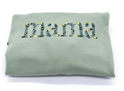 Detailansicht der Stickerei "Mama". Die einzelnen Buchstaben sind in Form von Blättern in verschiedenen Gruentoenen und gelben Blüten bestickt.